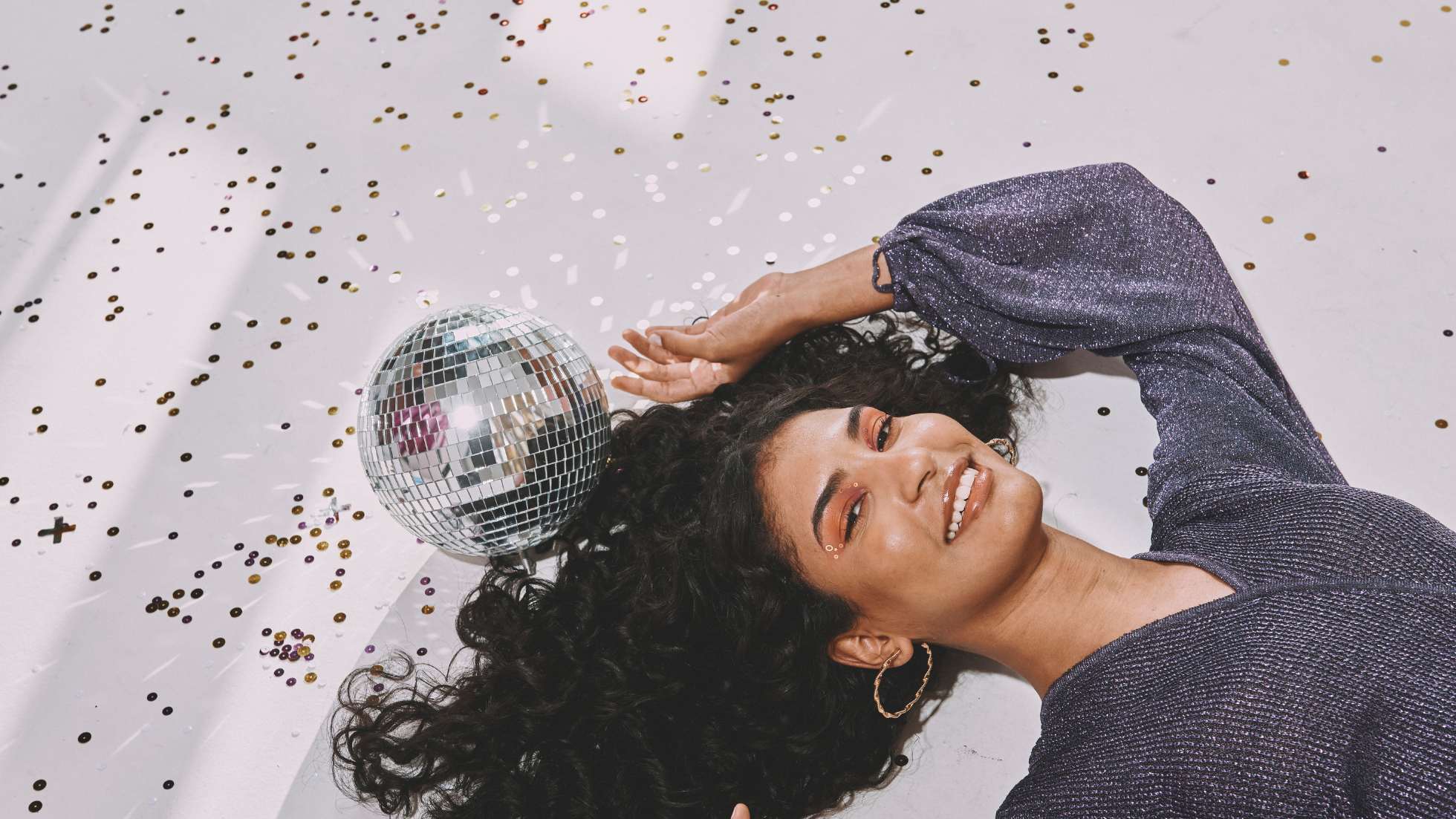 A woman laying on the floor next to a disco ball and glitter, looking up and smiling.