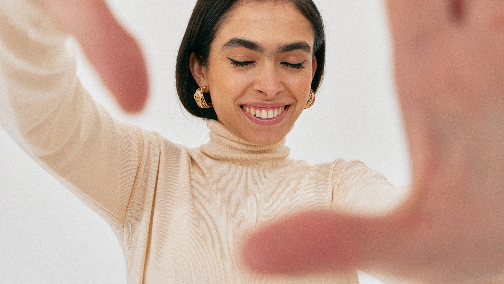 A woman holding how her hands towards the camera to frame her face while she smiles with her eyes closed.