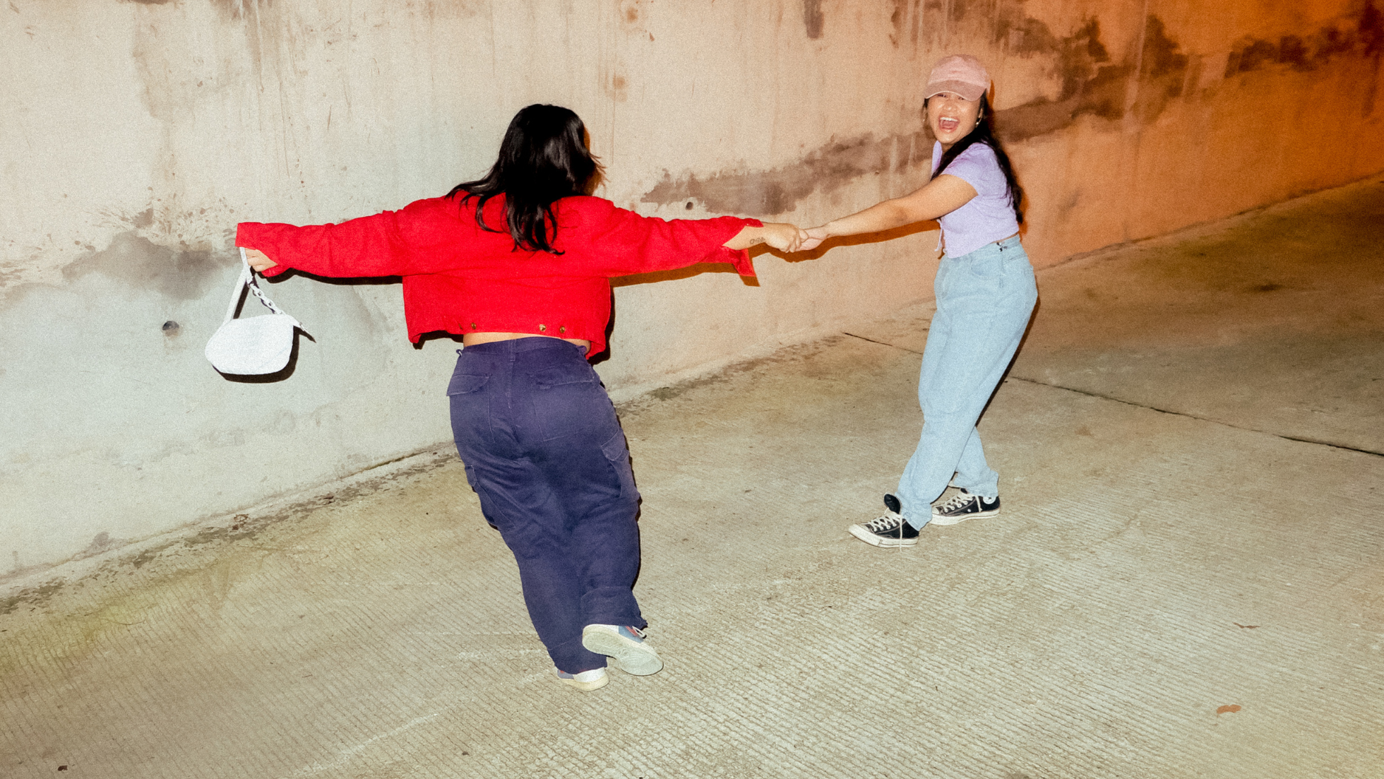 Two friends holding hands, swinging around on a sidewalk, laughing.